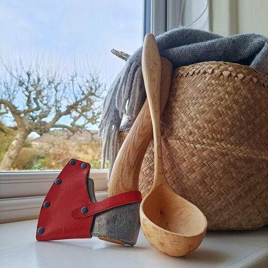 A hand carved bent beech branch ladle leaning on a woven basket, accompanied by a Kalthoff small carving axe. Apple tree in the background outside of the window