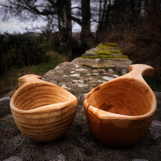Two sami kuksa drinking cups sitting on an old flat topped stone wall and with a pine forest background