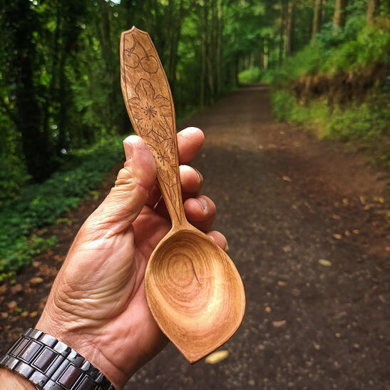A cherry spoon rests in the hand with a pathed woodland in the background. The spoon has a detailed cherry blossom design kolrosed onto the handle to further embellish the story of the spoons origin