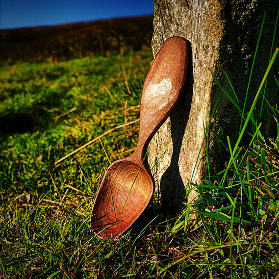 A baked alder symmetrical eating spoon rests in the golden sunlight against the base of a stone wall with a beautiful summer meadow as a backdrop