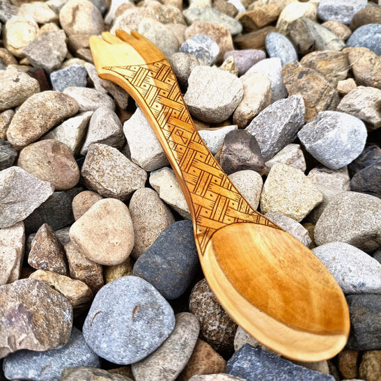 A basket weave kolrosed design on an apple wood spork, resting gently on the pebbles at the rear of a beach