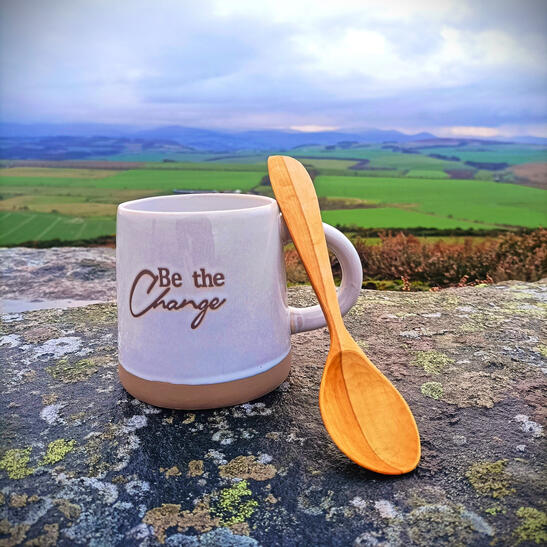 A hand carved two tone apple wood eating spoon resting on an oversized mug marked with be the change. Backdrop is a beautiful scenic view of the cheviot hills, vibrant colours
