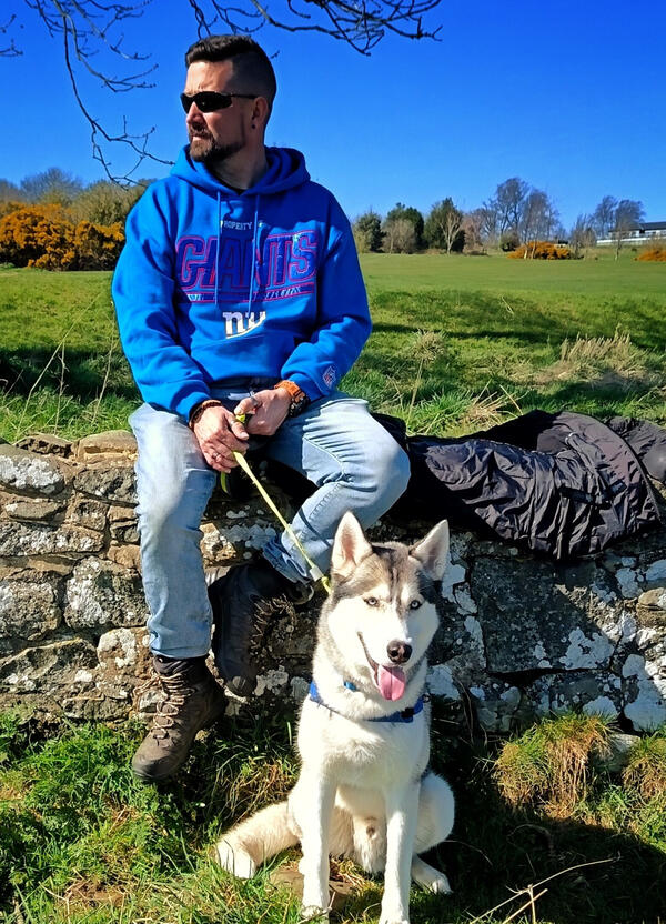 Nick sitting on a wooden bench by a stone wall looking into the distance with content