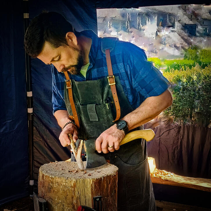 The maker Nick working on piece in his workspace, standing at the chopping block with axe, green canvas apron and blue checked shirt