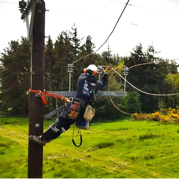 The maker Nick hangs on the side of a powerline pole, reaching to affix new wires to a new dark brown pole. Behind a little softwood copse and vibrant grass field