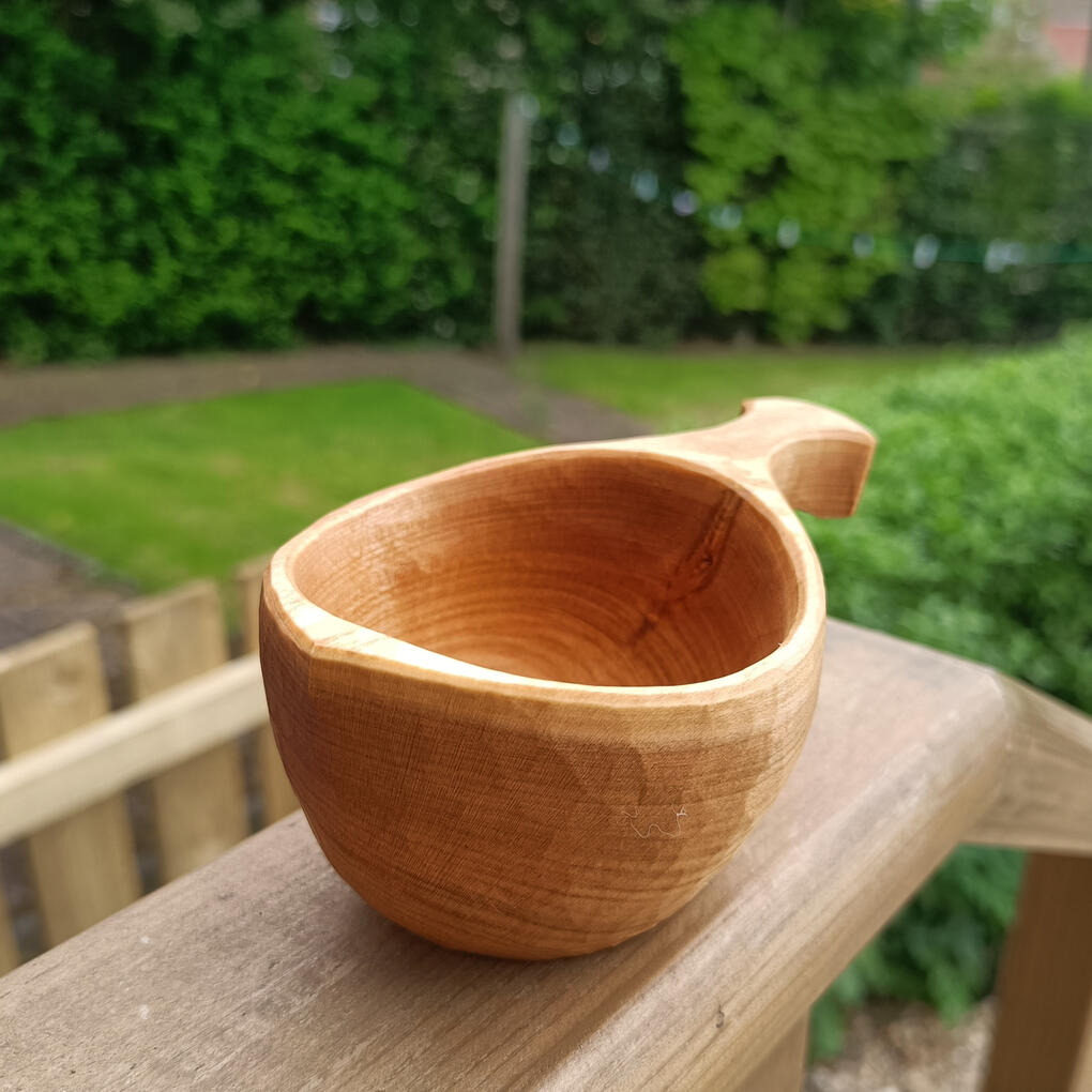 a small cherry sami kuksa cup shot on a wooden garden decking rail with a view of the lawn and dark beech hedge at the rear